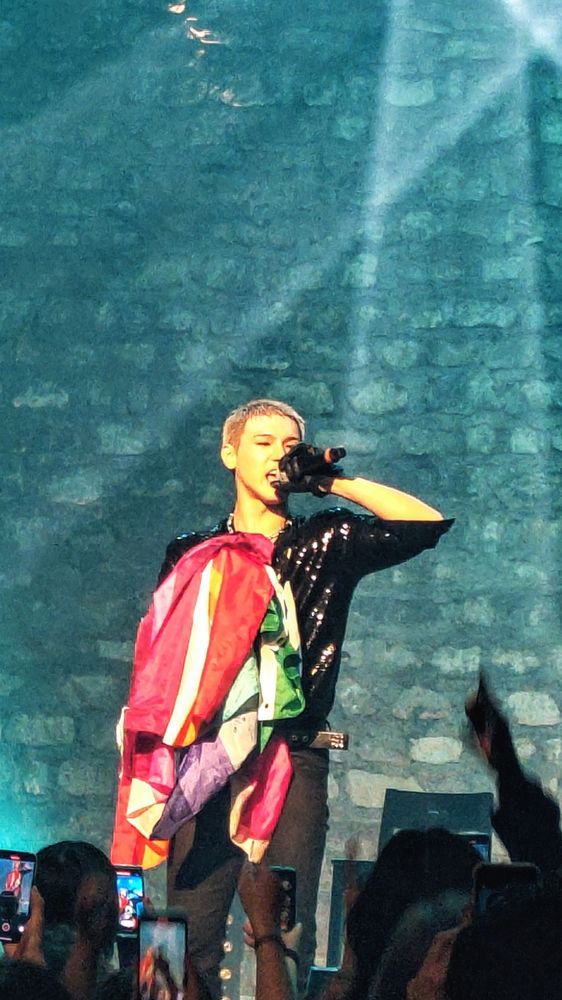 BAIN sur la scène du Café de la Danse le jeudi 16 octobre 2025. Chemise noire à paillettes, pantalon et gants noirs. Il chante en portant de nombreux drapeaux LGBTQIA+.