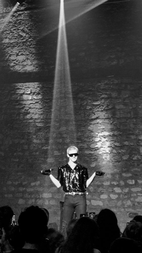 BAIN sur la scène du Café de la Danse le jeudi 16 octobre 2025. Noir et blanc. Chemise noire à paillettes, lunettes de soleil, pantalon et gants noirs.