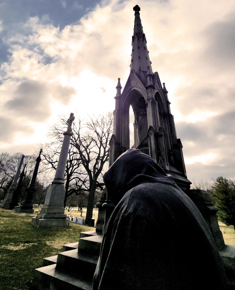 Photo of a hooded figure from the side, in front of a large gothic styled memorial marker. Taken at Oakwood Cemetery in Joliet, Illinois. A row of memorial obelisks taper off into the distance.