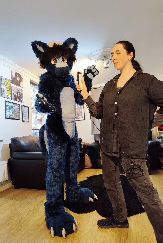 Navy blue dragon fursuit posing in a living room with a lass in black grinning with clippers in their hand implying they're going to shave the fursuit