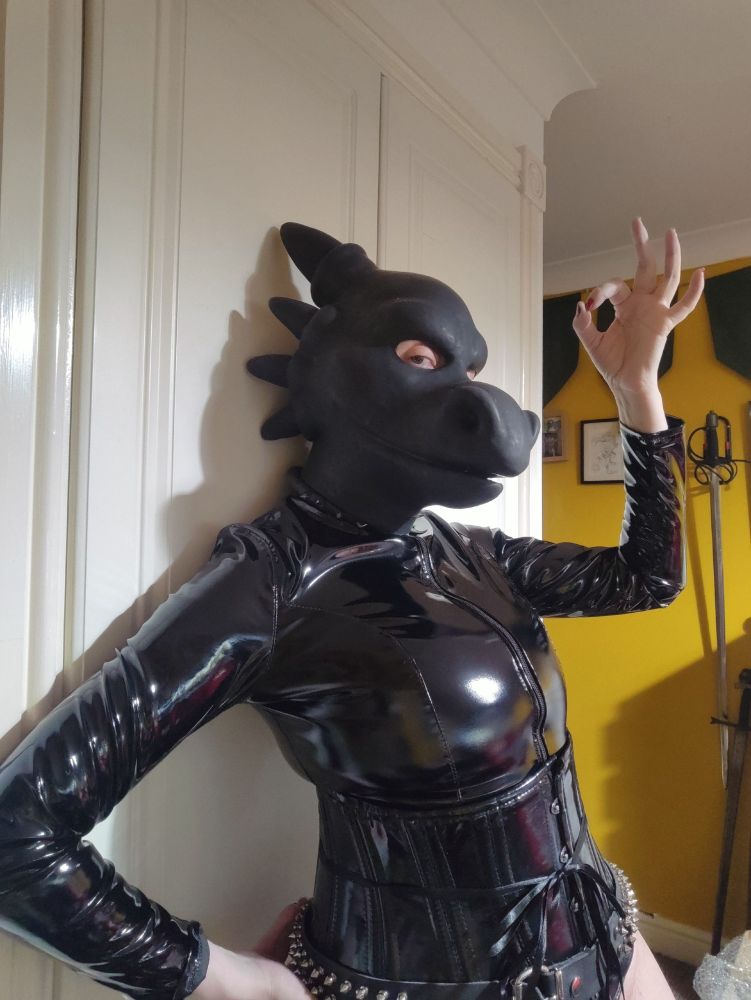 lass wearing a rubber dragon hood and a PVC top bodysuit with a PVC corset and a spiky  belt. Sword in the background
