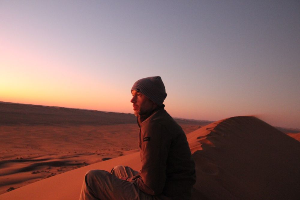 Dom sitting on a dune at Narabeb in a beanie, with wind blowing the sand and the sun setting. 