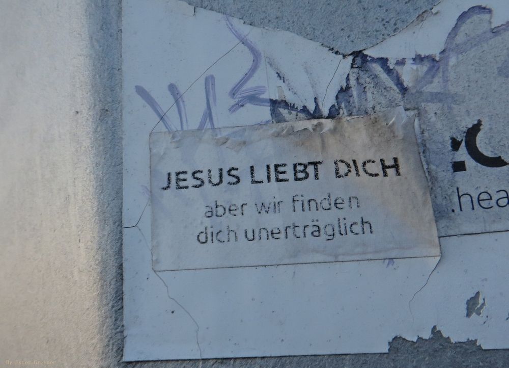 Werbetafel mit abgerissenen und zerschlissenen Plakaten. Daran klebt ein kleiner Sticker: Jesus liebt Dich
aber wir finden dich unerträglich.