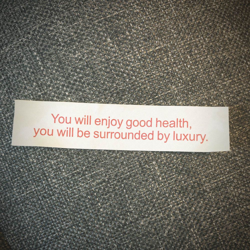 A white slip of paper from a fortune cookie with red text which reads “You will enjoy good health, you will be surrounded by luxury”