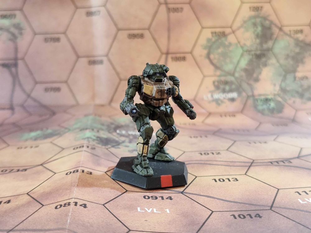 A Vindicator mech mini from the game Battletech painted in an olive drab, tan, and yellow color scheme. It is placed on a desert map sheet as a background.