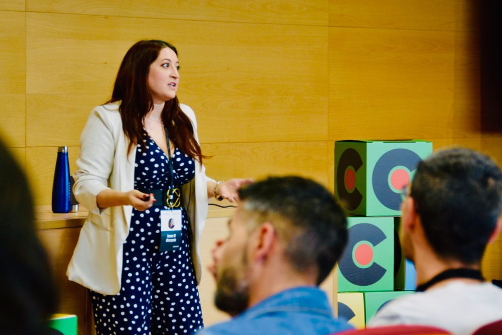 Fotografía tomada por Commit, en la que se ve a Irene en el escenario dle track, hablando al público. Lleva un traje con lunares y una americana de color claro, y se ve su acreditación. A su lado hay cubos con el logo de CommitConf y tras ella una  botella de agua apoyada en una mesa.