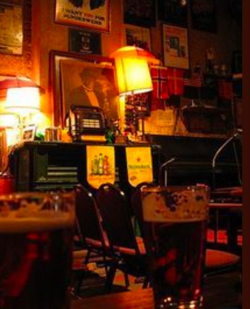 Photo of two pints of beer (partially drunk) in gloomy light against the backdrop of a pub wall with eclectic posters and framed portraits 
