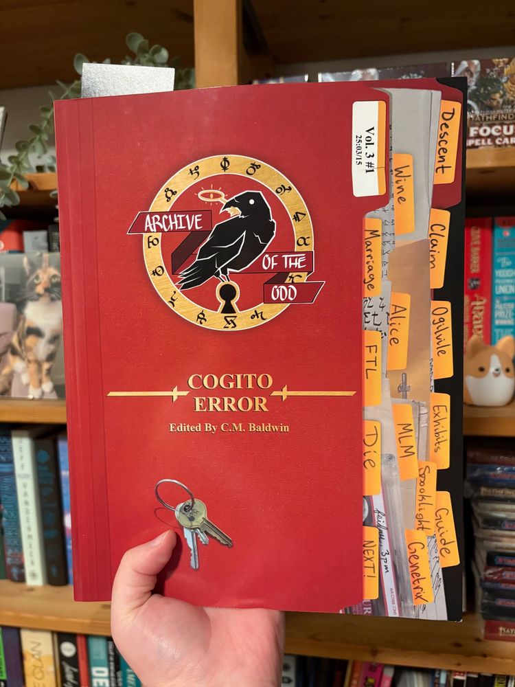 The zine held up in front of a bookshelf. It's a large softcover book, and the cover is stylized to look like a bunch of file folders.