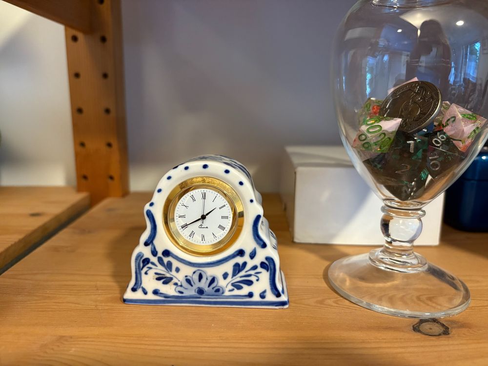 A little clock in ceramic stand, with blue floral highlights in the Delft style. Next to it is a glass jar with dice in it.