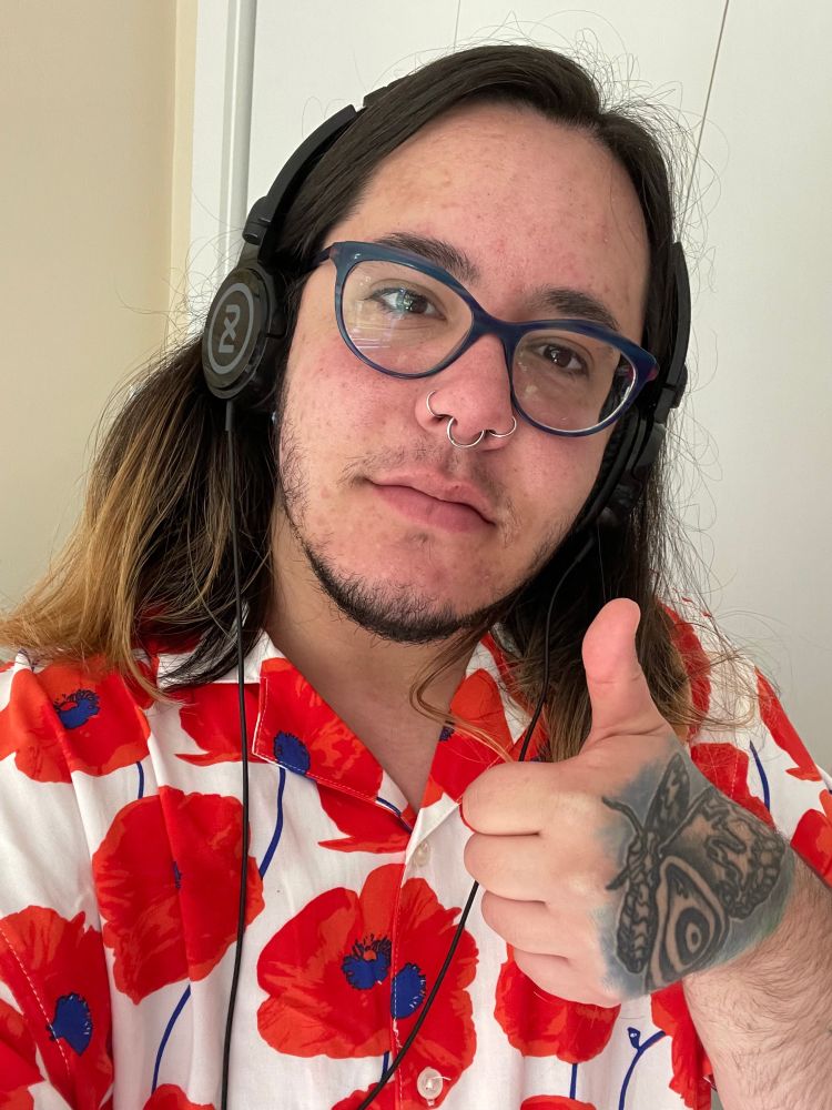A selfie of a light-skinned person wearing headphones and a shirt with large orange flowers, They are giving a thumbs up and have a tattoo of a moth on their hand.