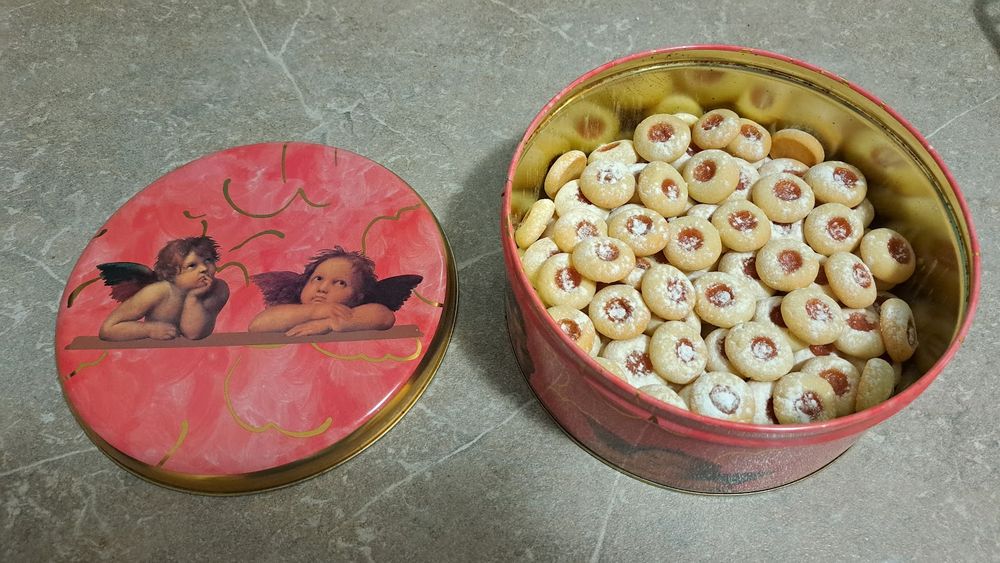 Eine runde Plätzchendose gefüllt mit runden Plätzchen mit Marmeladenfüllung und Puderzucker bestreut steht auf einer marmornen Anrichte. Daneben liegt der rosa Deckel, der zwei Engel zeigt, die sich auf einer Art Brett abstützen.