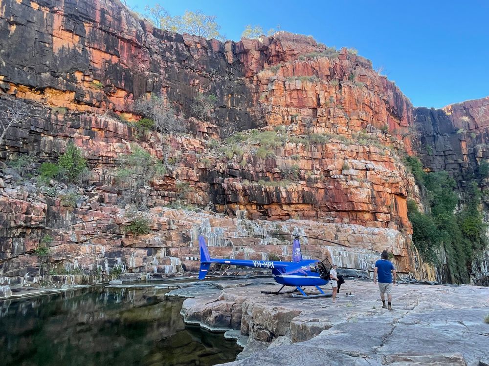 A blue helicopter landed on a flat rock in a gorge with a sheer orange rock face behind it, a tranquil water pool beside it and bright blue skies. The pilot and her passenger in the frame also 