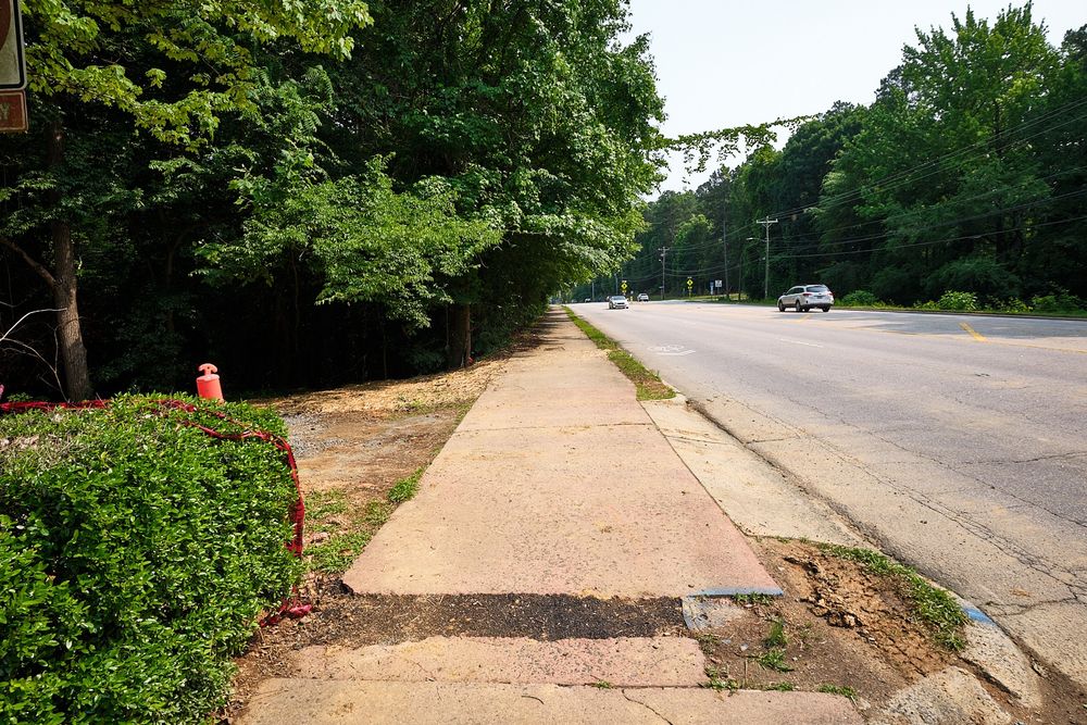 Rose colored sidewalk beside a 4-lane highway with a hole stretching across the width of the sidewalk filled with some type of asphalt composite and previous long cable stretching across the sidewalk from the right side, now removed.