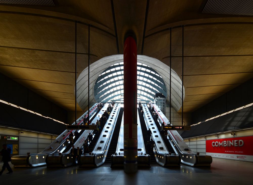 Underground rail station at Canary Wharf showing two sets of escalators going up and down; a pole in the foreground center and escalators seem to be emptying into a windowed dome above.
