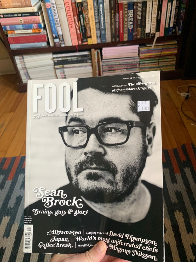 A copy of Fool Magazine, issue 3, held in the foreground. American chef Sean Brock is on the cover. In the background, à bookshelf full of magazines and books. 