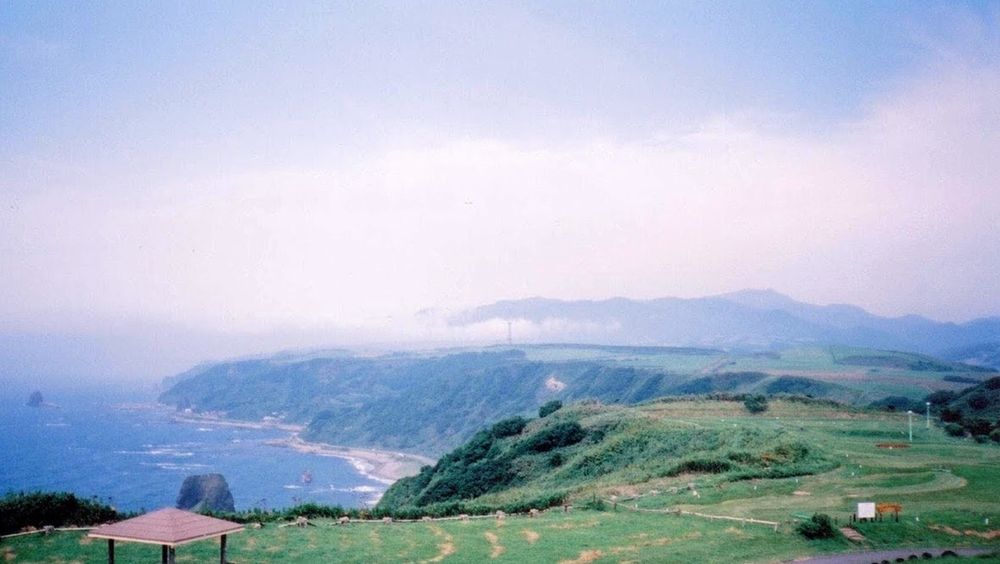 北海道・立象山展望台から撮影された広大な海岸線の風景。手前には芝生の広場が広がり、左下に屋根付きの東屋の一部が見える。海岸線は深く緑の木々に覆われた崖が続き、崖の下には青い海と岩礁が点在している。遠景の山々は低い雲や霧に覆われ、空は白くぼやけている。全体的に霧がかったような、柔らかなトーンの景色。