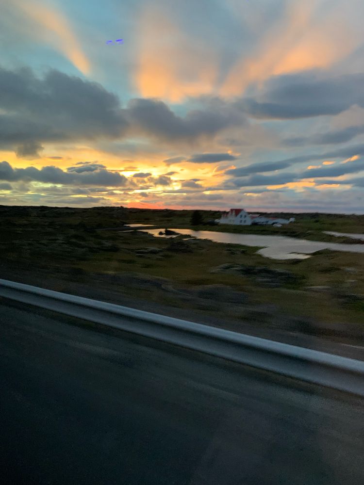 Roadside view of sunset on the way to Keflavik. Sky is littered with tufts of clouds magnificently lit by sun in yellow, pink/purple and orange. Towards the horizon is a singular building with some water mirroring the sky in front of it.
