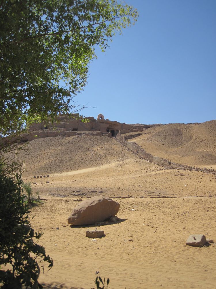 Blick über sandigen Boden auf eine Treppe, die einen kargen, braunen Hügel hinaufführt. Auf dem Hügel sind Ruinen zu erkennen. Im Vordergrund ragt ein grüner Baum ins Bild. Der Himmel ist strahlend blau.