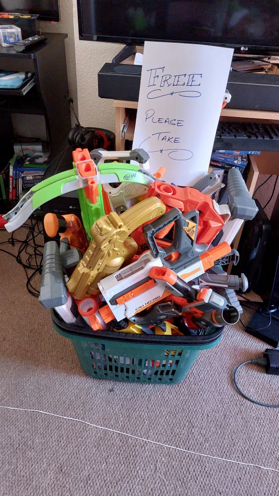 A basket full with netf guns, about 11 visible, 16 total. Has a handwritten sign - free please take