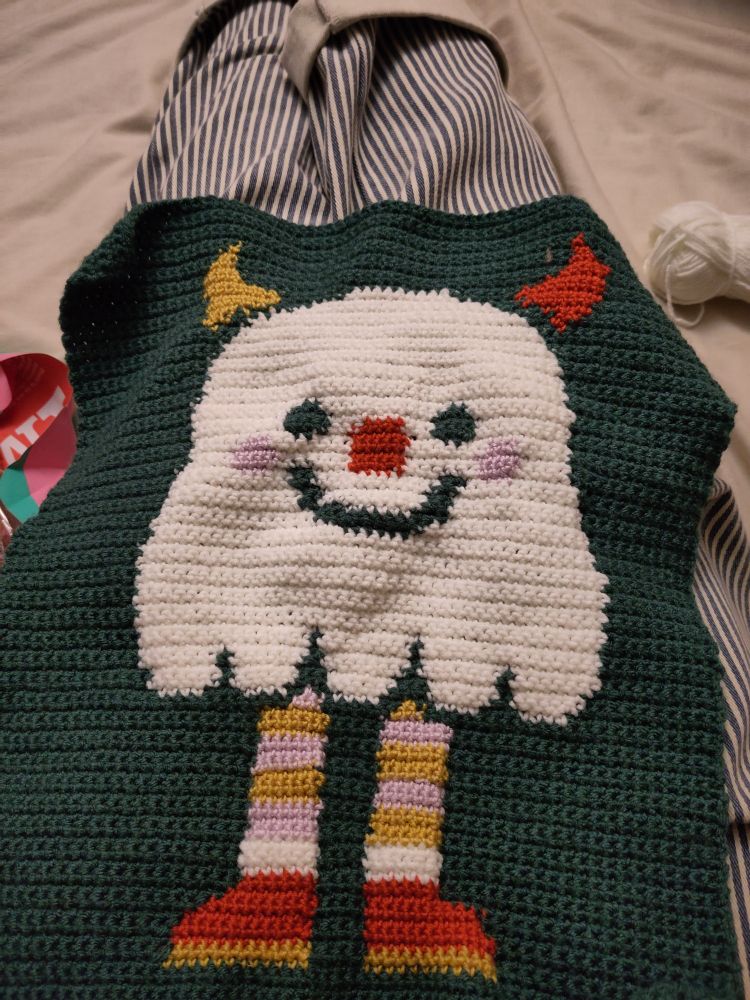 A picture of a crochet tapestry with a dark green background and a white ghost. The ghost is smiling and has horns, stripey stockings and red shoes.