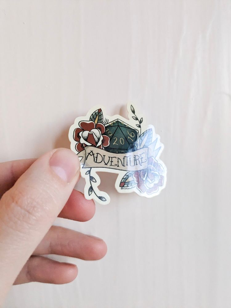 A picture of a hand holding up a sticker of a D20 dice surrounded by flowers and a banner that says "Adventure." It's landed on a nat 20 and the sticker is in the style of a traditional color tattoo.