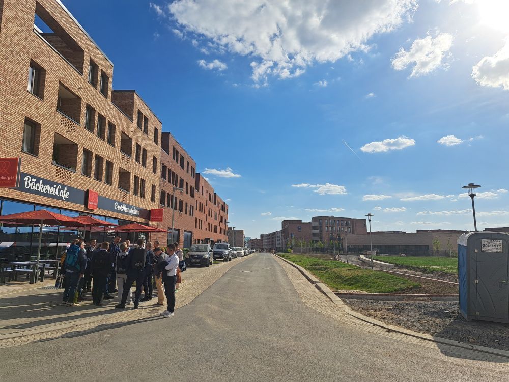 Die Exkursionsgruppe steht an einer Kreuzung im Neubaugebiet. Links und im Hintergrund sind bereits Klinkerbauten fertiggestellt, rechts ist noch Grün- und Brachfläche, die zu einer angrenzenden Schule gehört.