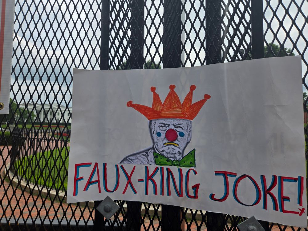 Protest sign w Trump as a clown wearing a crown and it says FAUX KING JOKE! Sign is placed on the security fence with white house far in the backround
