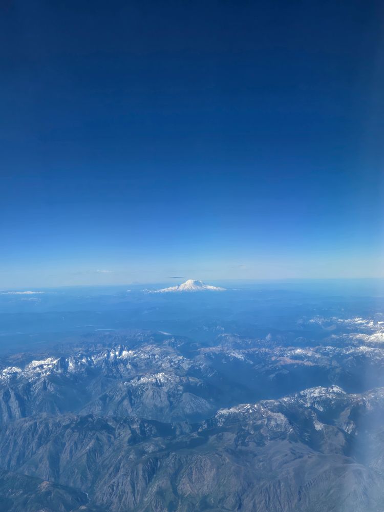 Mount Rainier, as seen from a plane, rises in the midst of the cascade mountain range. Rainier is white and snow capped but surrounded by shades of blue on land and in the bluebird blue sky