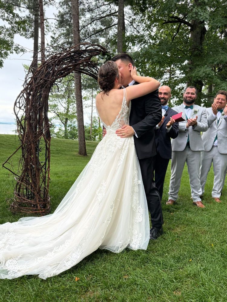 A man in a black tuxedo kisses a woman in a white wedding dress on a lawn under trees. Groomsmen and an officiant clap in the background