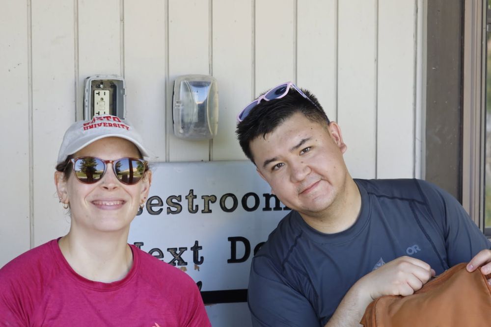 A man and woman smile at the camera in front of a beige wall