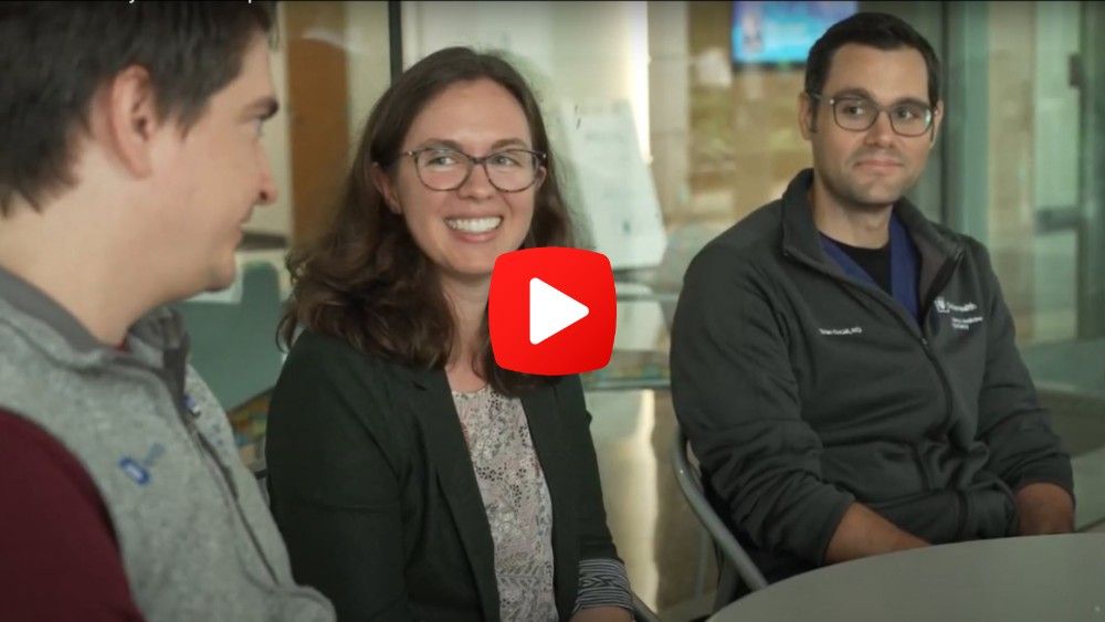 Three adults talking and smiling while sitting at a table. "Play" button is overlaid on image.