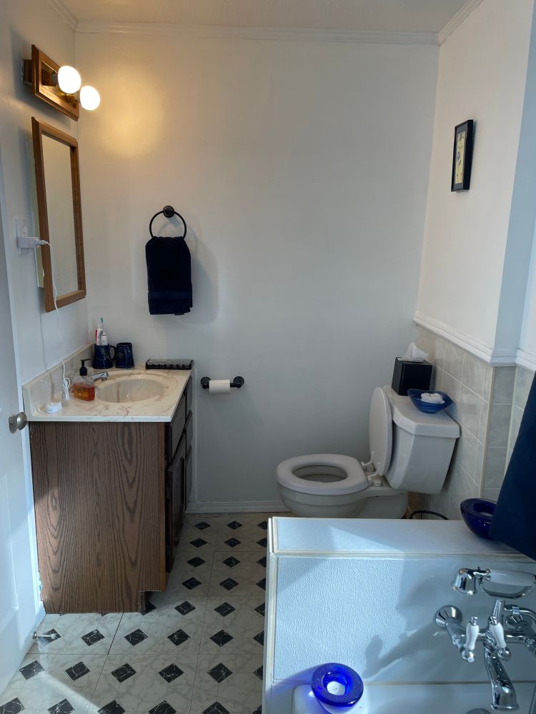 Other half of a small bathroom where it gets worse, with a dark brown vanity under a cream and brown marbled sink, with medium brown mirror and light fixture above. One wall is the marbled gray tile, and the floor is a white and black marbled diamond pattern, which is just truly over the top.