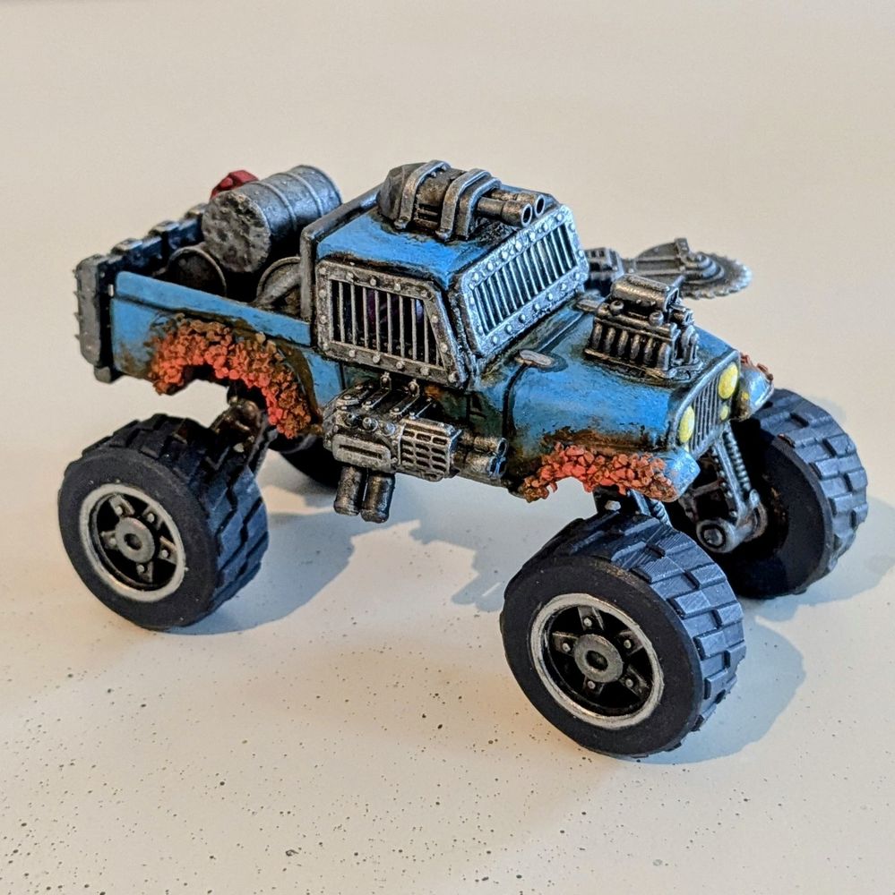 A rusted out monster truck with cannon turrets and a large buzzsaw coming out of the side. It has an exposed hotrod engine, oil barrels in the truck bed and bars over the windows 