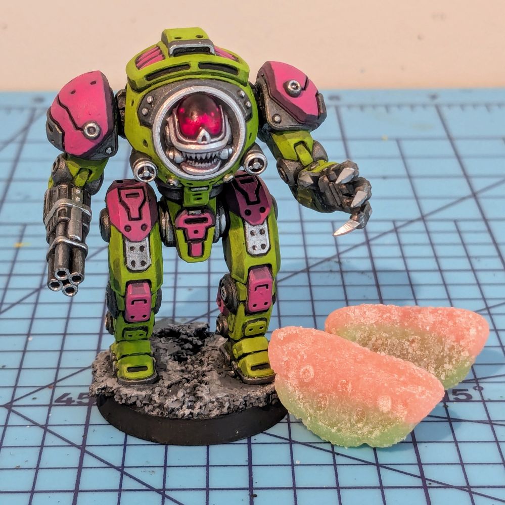 A mech miniature standing next to two watermelon candies. The mech has a claw hand and a cannon hand and a creepy skull-like face 