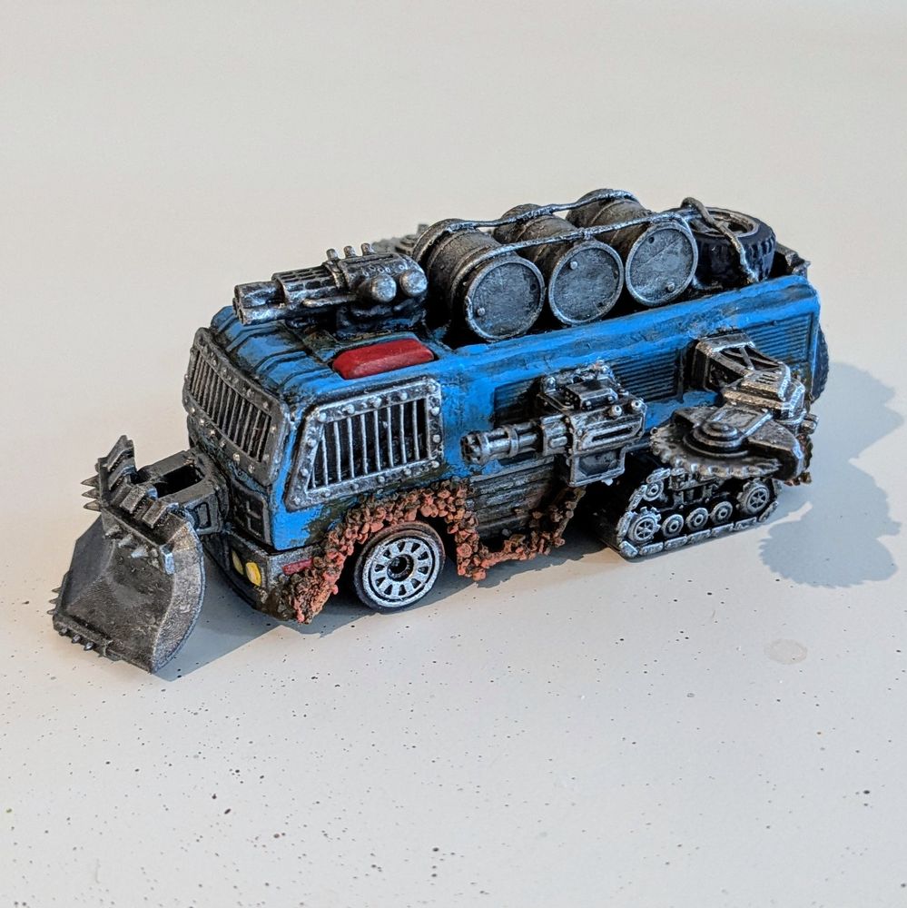 A rusted out bus with a spiked plow on the front, barred windows, gun and saw turrets, and three oil barrels strapped to the roof 