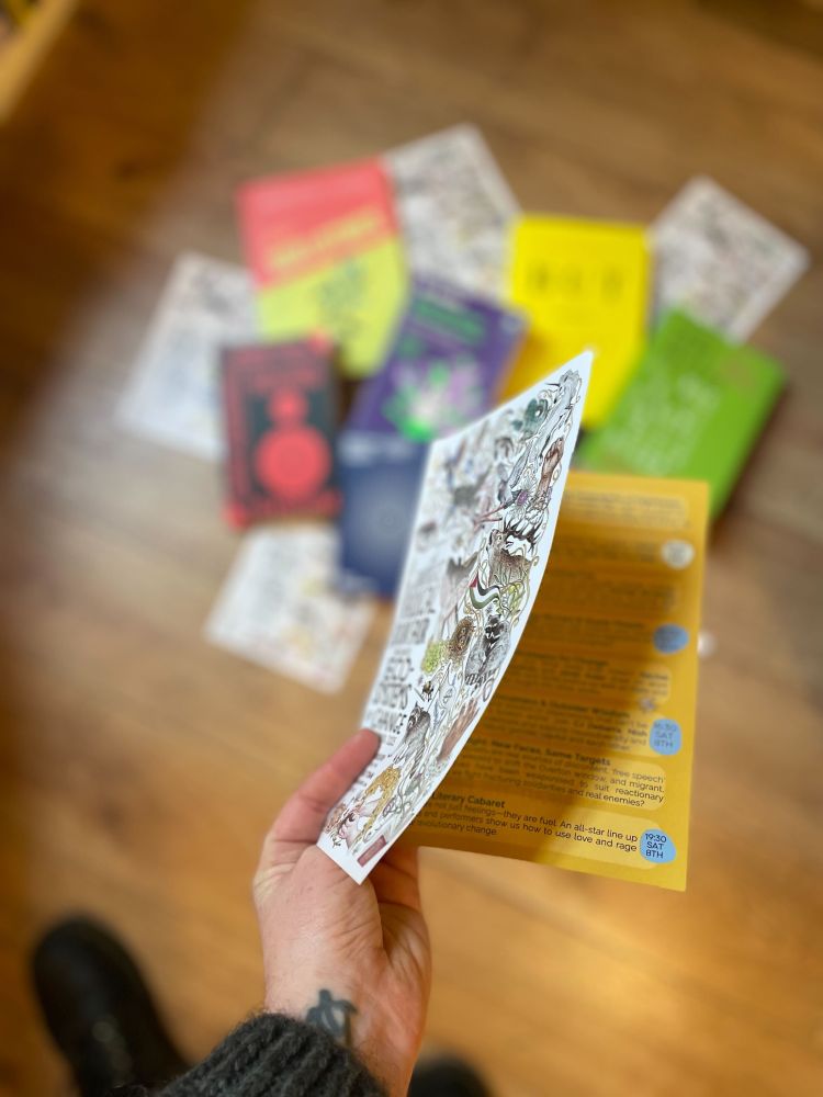 Slightly different angle on the first picture where a white hand with a wee tattoo holds up a leaflet with a richly illustrated cover full of racoons and mythical creatures and flags and books and the text reads Edinburgh's Radical Book Fair, there is just a peek of some of the events listed inside.