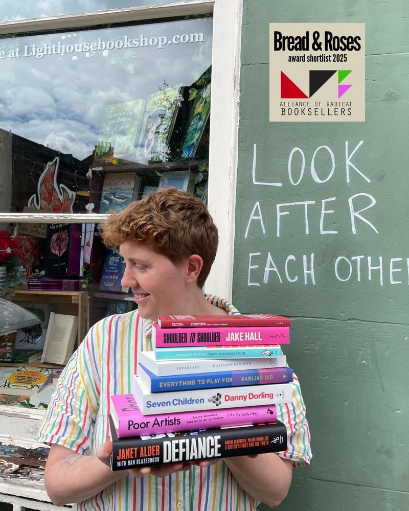 In front of Lighthouse Bookshop, which has a green wall on which is written "Look after each other", stands Mairi, Lighthouse's owner. Mairi has short ginger hair and is wearing a stripy top, and Mairi is holding the shortlisted books for the Bread and Roses Award: Pleasure Gardens, Shoulder to Shoulder, Race and Education, Intervals, Everything to Play For, Seven Children, Poor Artists and Defiance. In the upper right-hand corner is the logo for the Alliance of Radical Booksellers, which consists of black, red, green and purple triangles.