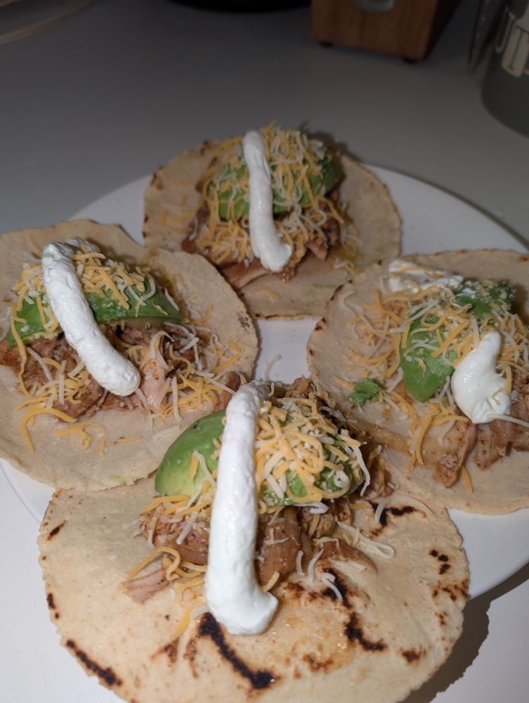 4 open faced Chicken tacos with sprinkled cheese, quartered avocado, and a thick strip of crema on grilled corn tortillas on a white plate.