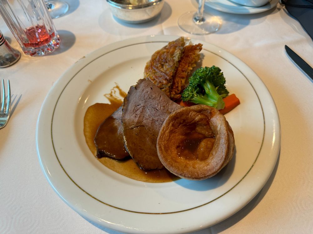 This image depicts a classic roast dinner plated with precision. The main component is slices of tender roast beef, served with a rich brown gravy. Accompanying the meat is a crispy Yorkshire pudding, a quintessential feature of a British roast meal. On the side are vibrant steamed vegetables, including broccoli and a carrot baton, adding freshness and color to the dish. Completing the plate is what appears to be a golden-brown potato rosti or another crispy potato accompaniment, providing a hearty and satisfying texture. The setting suggests a refined dining experience.