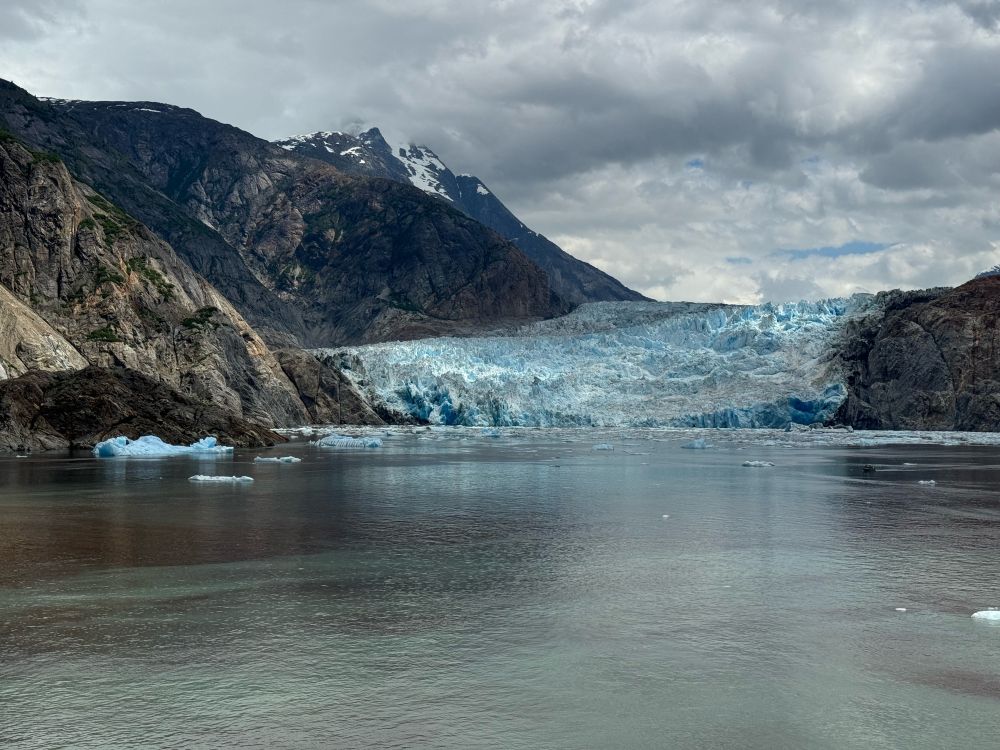 A snowy blue and white icey glacier nestled between rock formations. High snow capped mountains are visible in the distance.