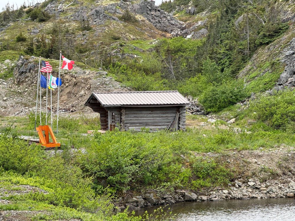 This image shows a small, rustic log cabin nestled in a lush, mountainous setting. The cabin has a simple, corrugated metal roof and is surrounded by greenery, with rocky outcroppings and trees in the background. In front of the cabin, a cluster of flagpoles display flags, including the American and Canadian flags, symbolising a cross-border historic location. A bright orange chair adds a pop of colour to the serene, natural scene, complemented by a small water body in the foreground. This tranquil spot reflects a sense of remote wilderness and history.
