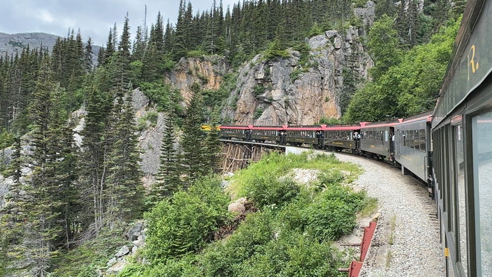 A scenic train ride through a lush, mountainous landscape. The train curves along a track surrounded by dense green pine trees and rocky cliffs, with parts of the track elevated on a wooden trestle bridge. The scene captures the natural beauty and engineering marvel of this rail route, emphasising adventure and exploration in the wilderness.