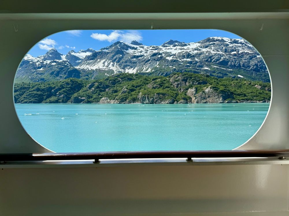 This image presents a picturesque view framed by the curved window of the ship. Through the window, a tranquil scene of turquoise water is visible, bordered by lush green hills and rugged snow-capped mountains in the distance. The clear blue sky above adds vibrancy to the landscape, while the framing creates a feeling of observing nature from a cozy, sheltered vantage point. The composition highlights the serenity of the natural setting.