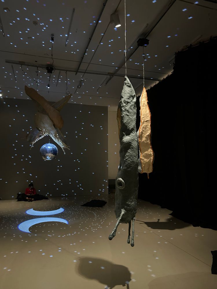 A shot of Abi Palmer’s Slime Mother exhibition showing some giant slug models hanging from the celling. One of the models if of two slugs intertwined above a disco ball that is throwing light all around the space.