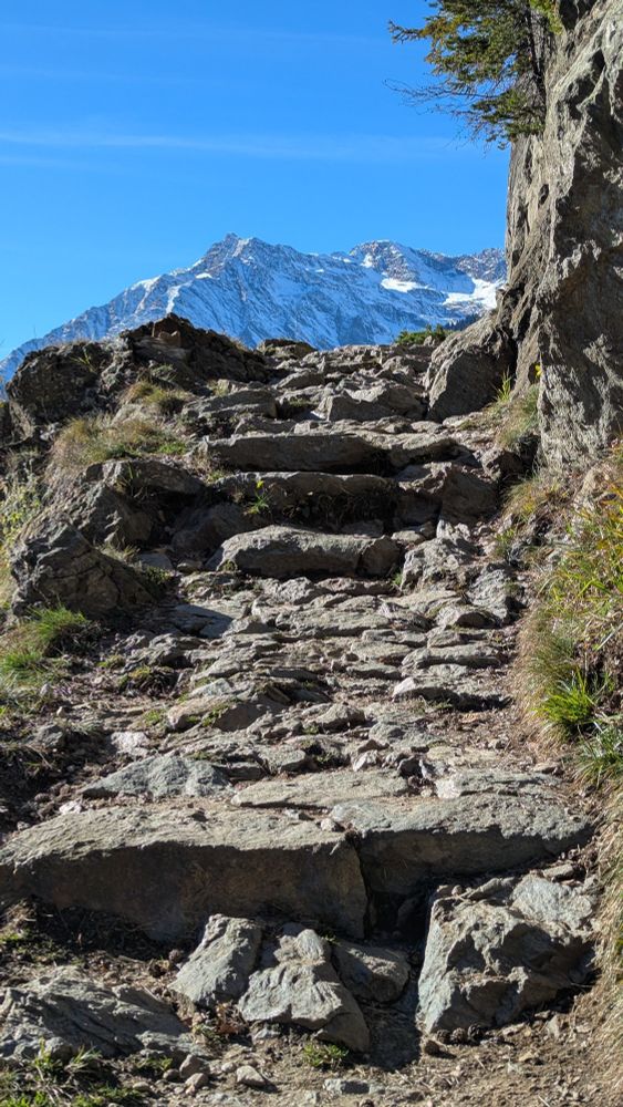 ein felsiger Weg mit groben, unregelmäßigen Steinstufen, im Hintergrund schneebedeckte Berggipfel 