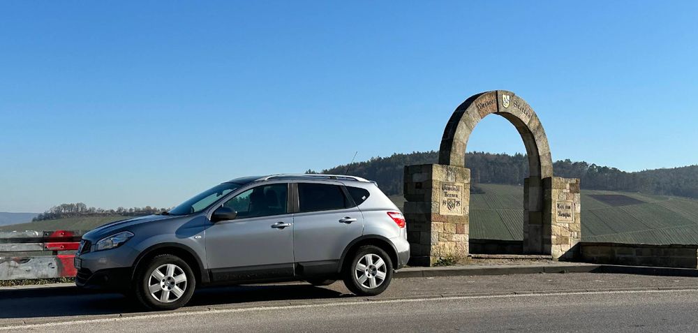 Screenshot eines Bildes der Kurve aus Google Maps vor dem Umbau. Rechts kann man ein ca. 3m hohes, freistehendes Steintor sehen. Links daneben steht in der Parkbucht ein silberner PKW.  Im Hintergrund liebliche Weinberge und Hügel.