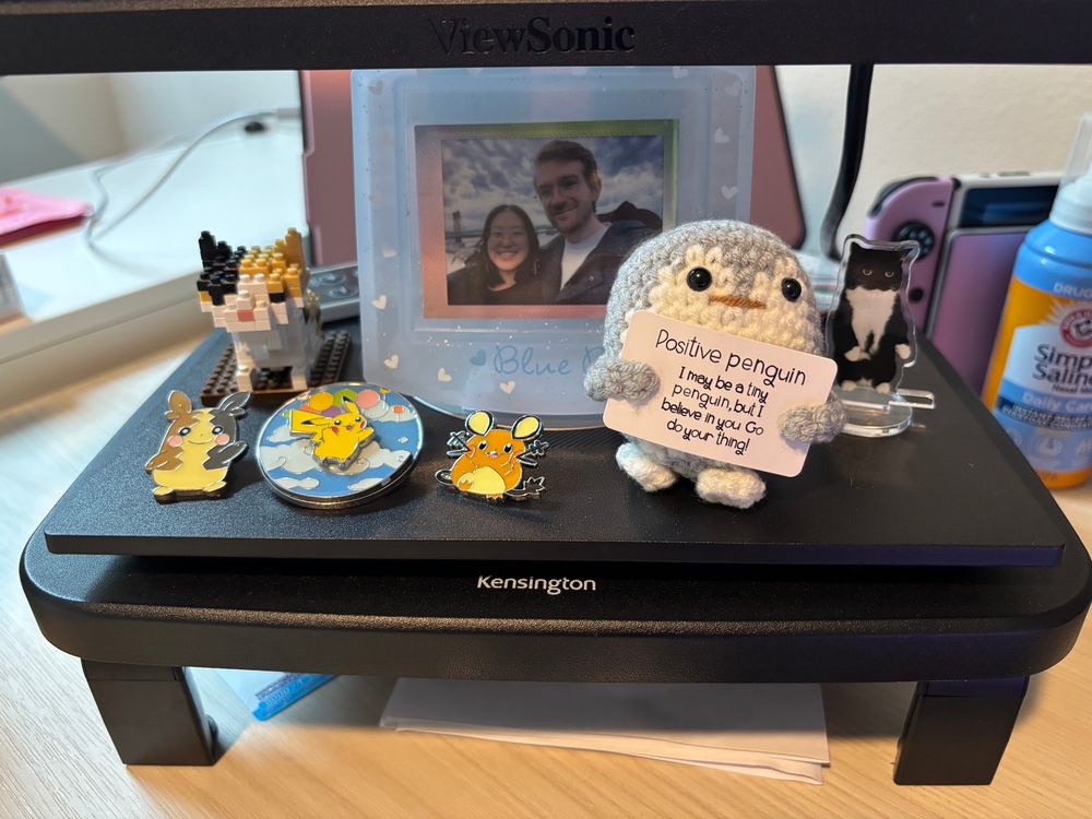 my little monitor riser with various things on it. cat lego, pokemon pins, a framed photo of nick and I, an uni the cat acrylic stand, and the newest addition is a crochet positive penguin. the penguin's sign: I may be a tiny penguin, but I believe in you. go do your thing!