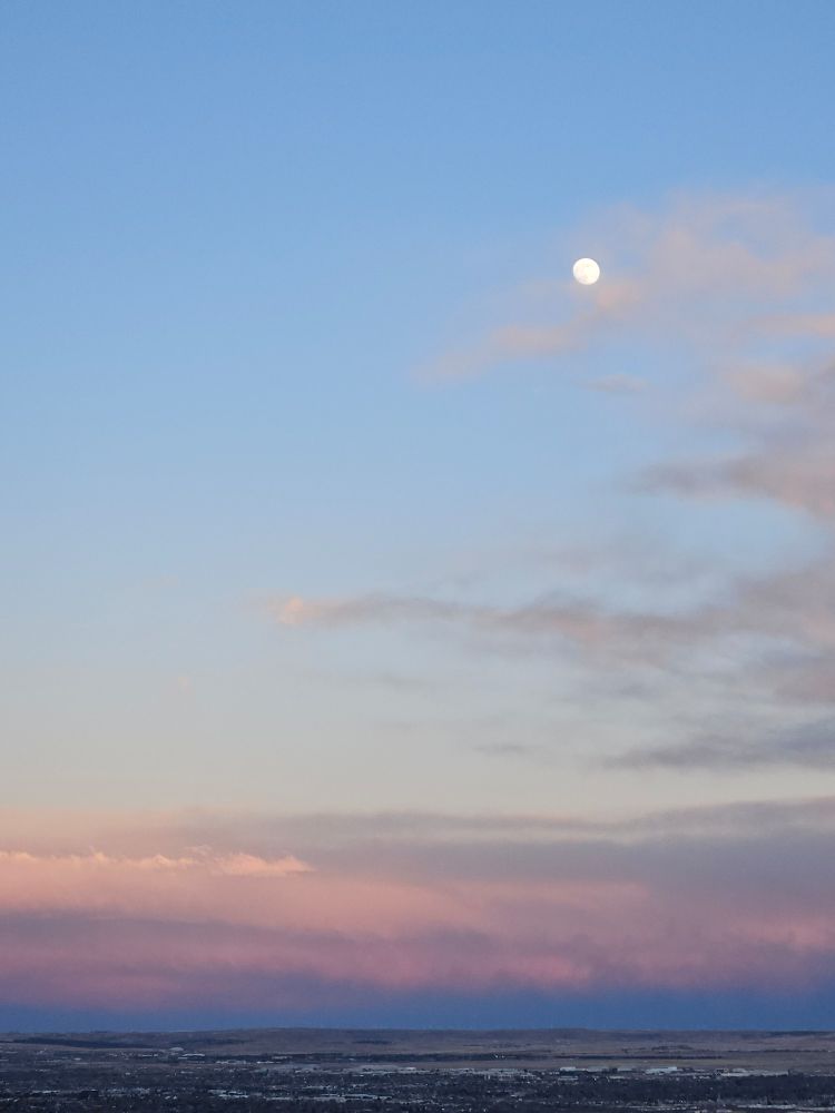 Tumultuous clouds low on the horizon at sunset with a nearly full moon high in the sky among more gentle clouds.