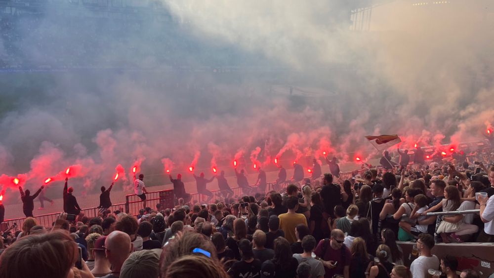 Blick aus der Gegengerade auf den Platz. Volle Ränge und Leute mit Pyro auf dem Zaun. Der Neben lässt den Blick auf die Tribünen dahinter verschwinden.