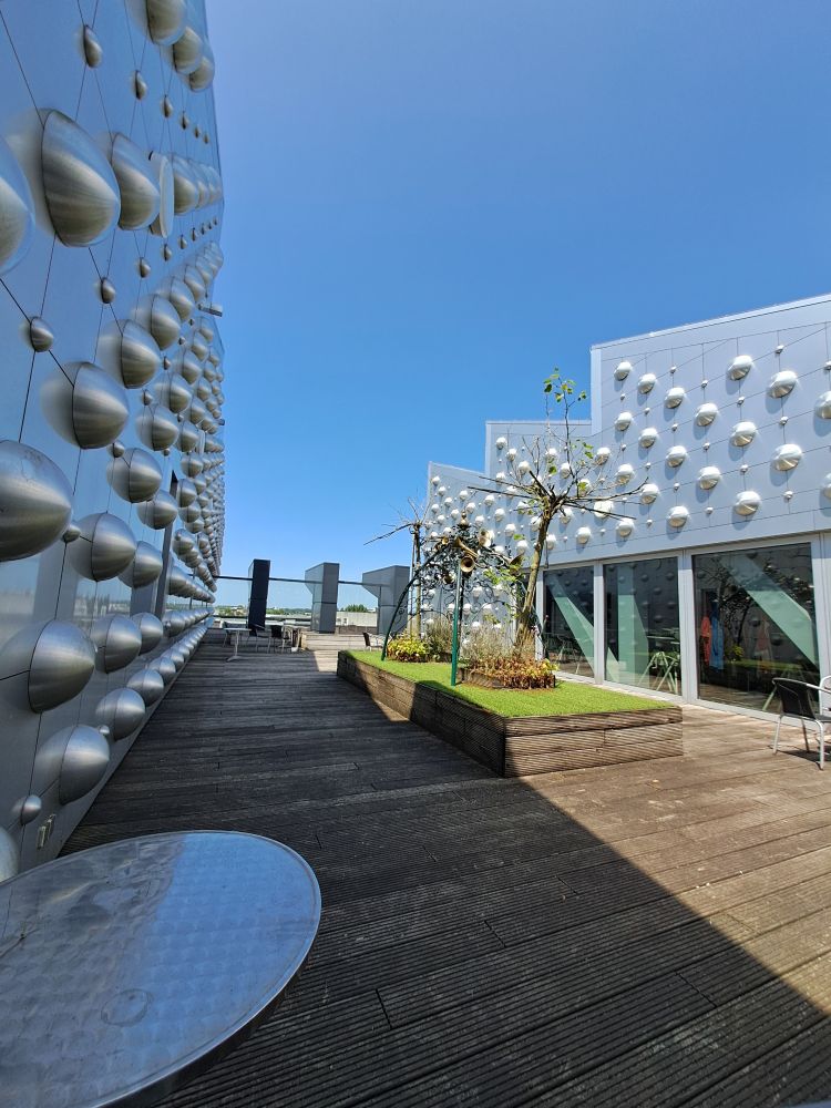 Links en rechts metaalachtige wanden met halvebolvormige uitsteeksels. Daartussen een houten 'terras' met in het midden een verhoging met (kunst?)gras en sprieterige boompjes. Tussen de boompjes een tentachtige metalen constructie met blaasinstrumenten erop.